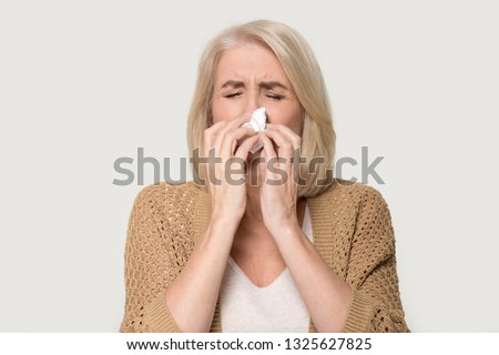 Ill allergic old mature woman blowing runny nose got hay fever rhinitis allergy flu, sick middle aged senior lady sneezing in tissue holding handkerchief isolated on white grey studio background Royalty-Free Stock Photo #1325627825