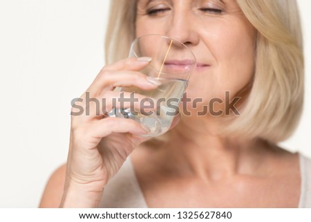 Healthy thirsty senior old woman drinking filtered pure fresh clean water, middle aged mature lady holding glass hydrating body isolated on white grey studio background, dehydration hydration concept