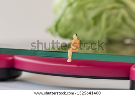 Miniature woman figure siting on the digital electronic bathroom scale for weight of human body. Fresh vegetables at shallow depth of field background. Healthy lifestyles and slimming concept.