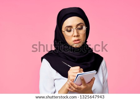    arab woman in black burqa is writing in a notebook on a pink background                           