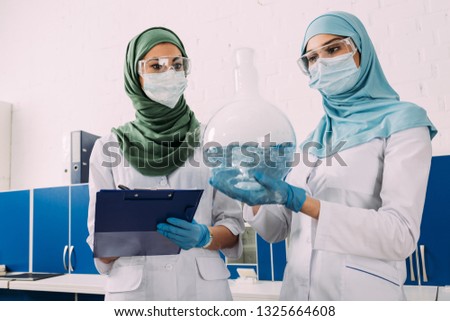 female muslim scientists holding clipboard and flask wth liquid in chemical laboratory