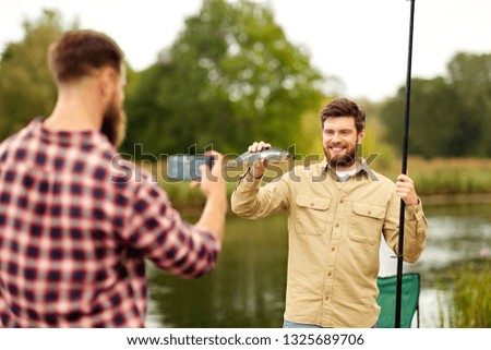 fishing, leisure and people concept - friend photographing fisherman with fish by smartphone at lake