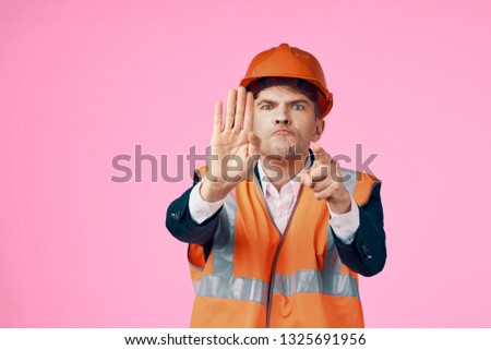 An engineer in orange helmets gesticulates with his hands on a pink background and a reflective vest                         