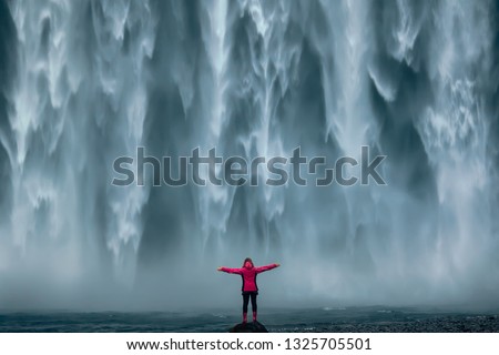 Iceland landscape photo of brave girl who proudly standing with his arms raised in front of water wall of mighty waterfall. Royalty-Free Stock Photo #1325705501