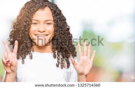 Young beautiful woman with curly hair wearing white t-shirt showing and pointing up with fingers number seven while smiling confident and happy.