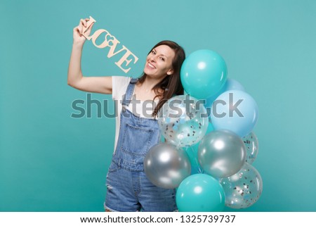 Cheerful young woman in denim clothes holding wooden word letters love, celebrating with colorful air balloons isolated on blue turquoise background. Birthday holiday party, people emotions concept