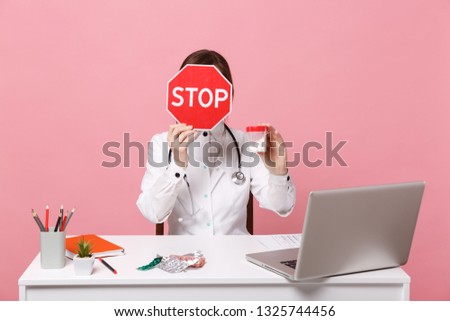 Female doctor sit at desk work on computer with medical document hold pills in hospital isolated on pastel pink background. Woman in medical gown glasses stethoscope. Healthcare medicine concept.