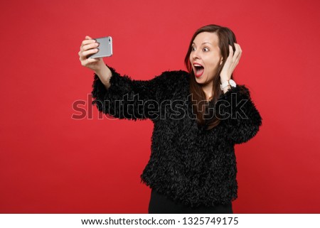 Excited young woman in black fur sweater clinging to head, doing selfie shot on mobile phone isolated on bright red wall background. People sincere emotions, lifestyle concept. Mock up copy space