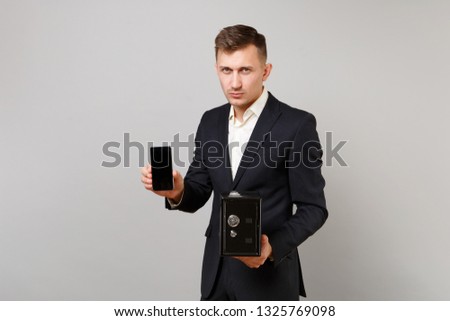 Serious young business man hold mobile phone with blank empty screen metal bank safe for money accumulation isolated on grey background. Achievement career wealth business concept. Mock up copy space
