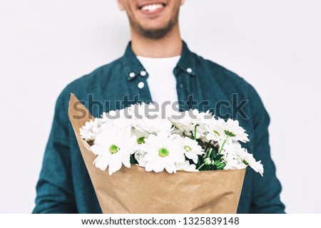 Cropped image of handsome male with a bunch of flowers preparing for a date with girlfriend. Young happy man smiling and delivering a bouquet of white flowers posing on white wall.
