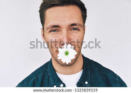 Closeup studio image of Caucasian handsome man smiling with a flower in mouth posing over white wall. Attractive male with a bunch of flowers preparing for a date with his girlfriend.