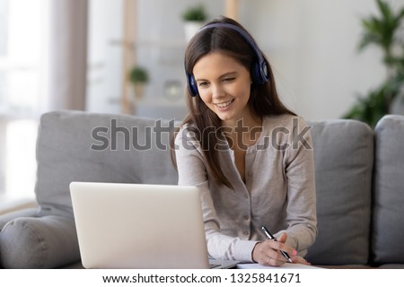 Smiling woman in headphones using laptop, writing notes, sitting on sofa at home, happy girl looking at computer screen, watching webinar, learning language online, distance education concept Royalty-Free Stock Photo #1325841671