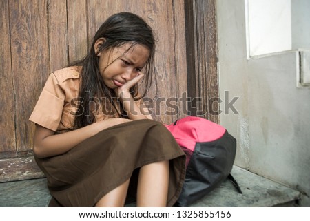 7 or 8 years child in school uniform sitting outdoors on the floor crying sad and depressed holding her backpack suffering bullying and abuse problem feeling alone and helpless as scared schoolgirl 