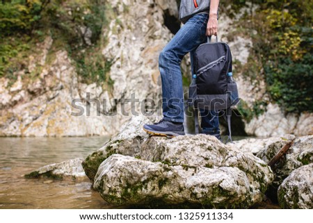 Unidentified male tourist in casual clothes is holding an equipped backpack for travel against the backdrop of a mountain river and stones. Concept of active recreation and comfortable clothing