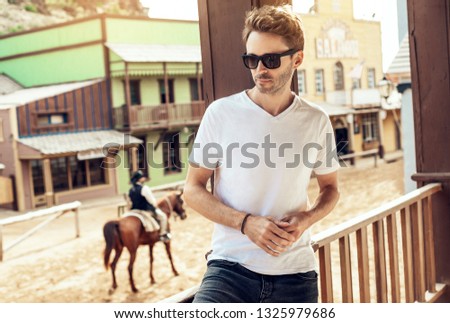 Young handsome bearded man is wearing a white empty blank t-shirt and standing on the western city street background. Horizontal mock-up style.