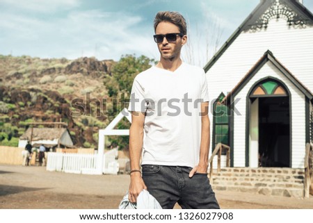 Young handsome bearded man is wearing a white empty blank t-shirt and standing on the western city street background. Horizontal mock-up style.