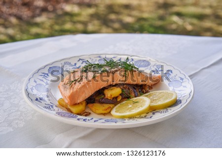  Close up picture baked salmon fillet with roasted potatoes, onion and slices of lemon and chopped fresh dill herbs served on rustic plate outdoor in garden restaurant during sunny spring day        