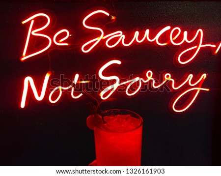 The glowing text ‘’Be Saucey Not Sorry’’ on the black background with the glass of mock tail with cherry