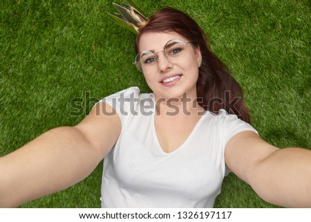 From above of adorable plus-size girl in white t-shirt and golden crown lying on green lawn taking selfie 