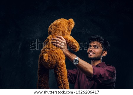 Young Indian guy in stylish shirt looks at his lovely teddy bear while holding it in hands. Studio photo against a dark textured wall