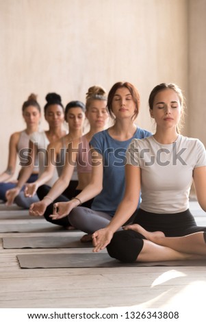Group of diverse young attractive people practicing yoga, doing Easy Sukhasana pose, working out indoor, mixed race female students meditating at sport club, yoga studio. Well being, wellness concept Royalty-Free Stock Photo #1326343808