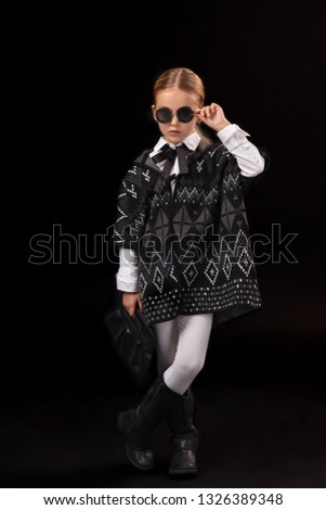 Little girl in a black glasses, dark poncho, black boots and a clutch in her hand