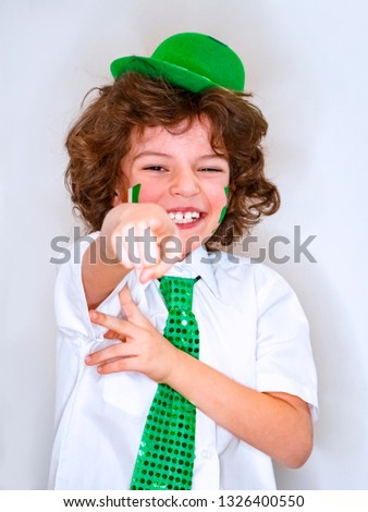 Curly child boy having fun during Saint Patrick celebrations over a light background.I am smiling boy with a Irish flag on my cheek pointing a finger at the camera