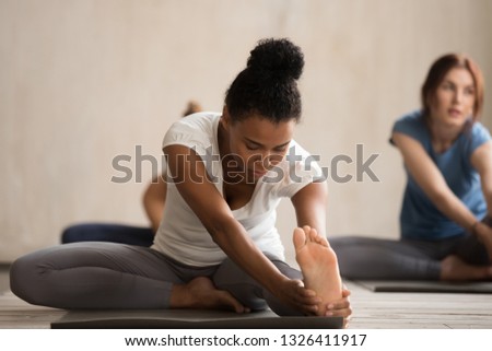 Yogi African woman and diverse group of young people doing yoga Head to Knee Forward Bend exercise, Janu Sirsasana pose, students working out indoor in club or studio. Well being, fitness concept