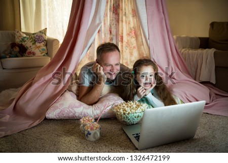 Family quality time. Father and daughter lie in homemade pink tent with flowers, watch cartoons on laptop, eating popcorn, laugh. Cozy stylish room. Family bonds concept 