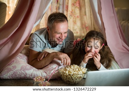 Family quality time. Father and daughter lie in homemade pink tent with flowers, watch cartoons on laptop, eating popcorn, laugh. Cozy stylish room. Family bonds concept 