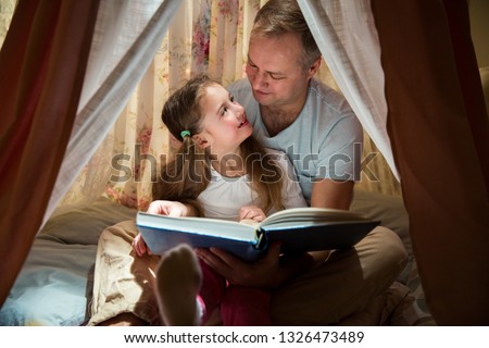Family quality time. Father and daughter sit in homemade pink tent with flowers, read big book, look at each other, smile and laugh. Cozy stylish room. Family bonds concept 