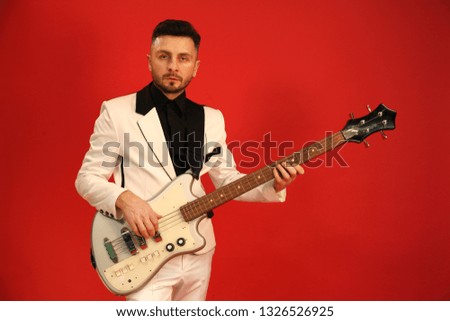 Handsome man in white classic costume with small beard playing bass guitar isolated on red background
