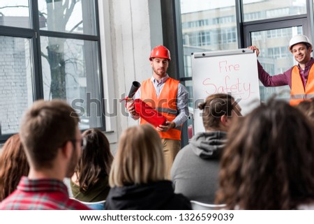 selective focus of handsome fireman in helmet holding extinguisher while standing near white board with fire safety lettering near coworker