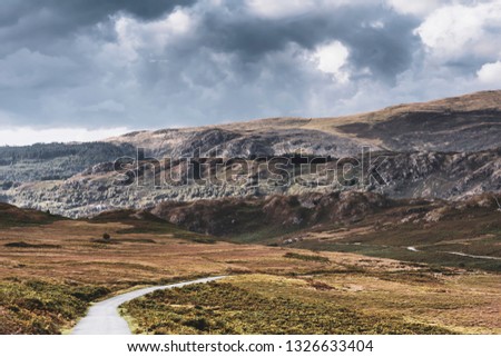 Country road with view on scenic valley and dark, dramatic sky above mountain range.Majestic landscape of Lake District,UK.Beautiful scenery of rural Cumbria.