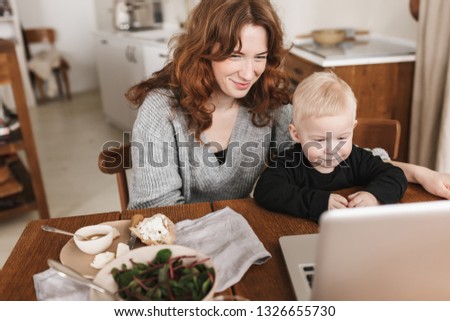 Young smiling woman with red hair in knitted sweater and little son sitting at the table with food and watching cartoons on laptop. Mom happily spending time with her baby in cozy kitchen at home