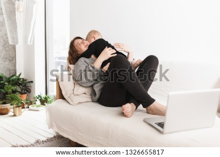 Young cheerful woman on sofa happily hugging her little son. Mom and baby boy dreamily spending time together with laptop in cozy living room at home