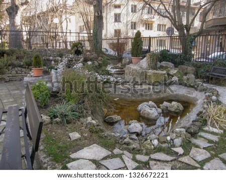 Patio with a small pond lined with stones, a bench and green bushes