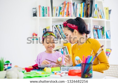 Picture of young mother teaching her daughter to paint Easter eggs at home