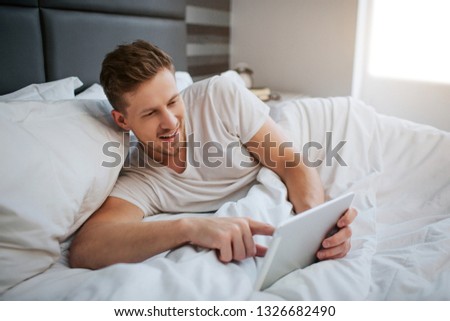 Young positive man lying in bed early morning. He hold white tablet and point on its screen. Relaxed. Daylight.