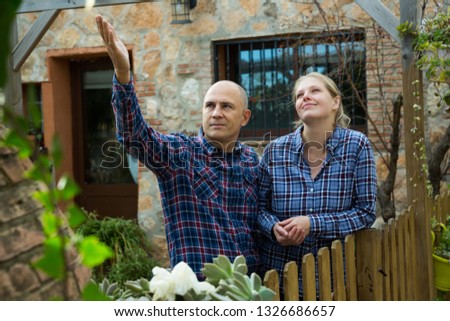 Portrait of positive man and woman standing together on courtyard of rural house and pointing to something