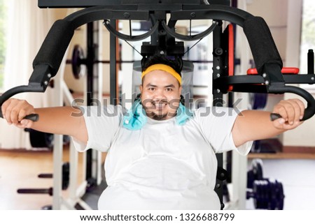 Picture of young fat man smiling at the camera while exercising with a fitness machine in the gym center