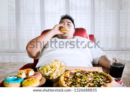 Picture of overeating fat man sitting on the sofa with junk foods in the living room
