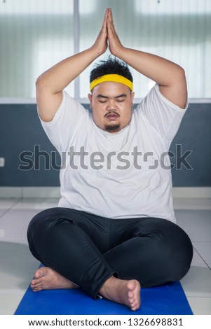 Picture of young fat man lifting hands while doing yoga exercise and sitting on the mat