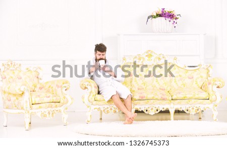 Elite leisure concept. Man with beard and mustache enjoys morning while sitting on luxury sofa. Man on sleepy face in bathrobe, drinks coffee, in luxury hotel in morning, white background.