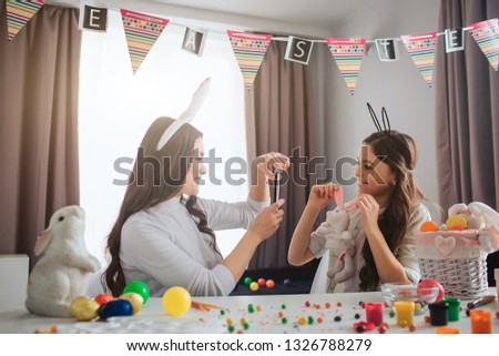Mother and daughter prepare for Easter. Young woman take picture of girl with rabbit toy. Girl pose. They wear easter bunny ears. People sit in room at table.