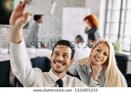 Happy business couple having fun at work and taking selfie with smart phone. Focus is on young businessman. 