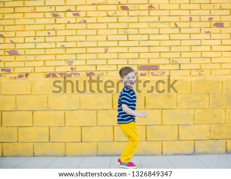 A five-year-old child in yellow pants and a striped T-shirt plays against the background of a yellow brick wall.