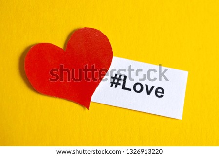 A popular hashtag "love" printed on white paper next to a red heart made of paper on a yellow background.
