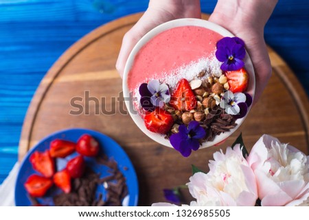 Young female hands holding pink smoothie bowl with strawberry, nuts, chocolate and decorated with flowers, top view. Spring healthy breakfast, overhead view.