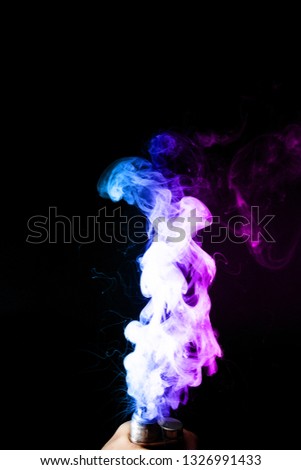 Young man holds vape in his hand. Vape clouds splash from vape coil on black background. Fog is dual color — blue and purple. Stock isolated colorful smoke with spray boiling glycerine.